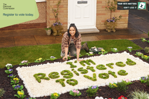 A woman kneeling by a flower bed with plants arranged to say 'I'm registered to vote'